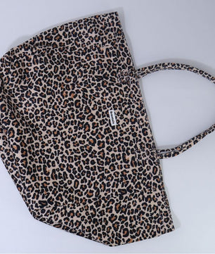 A leopard print tote bag with a beige, brown, and black animal pattern, featuring a single shoulder strap and a small white brand tag on the side, photographed on a light gray background.