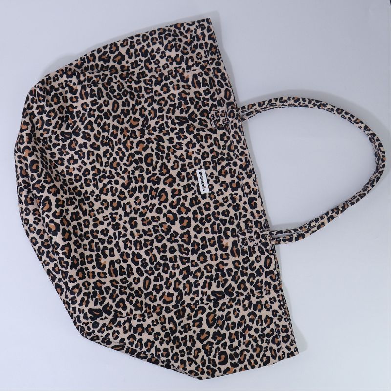 A leopard print tote bag with a beige, brown, and black animal pattern, featuring a single shoulder strap and a small white brand tag on the side, photographed on a light gray background.