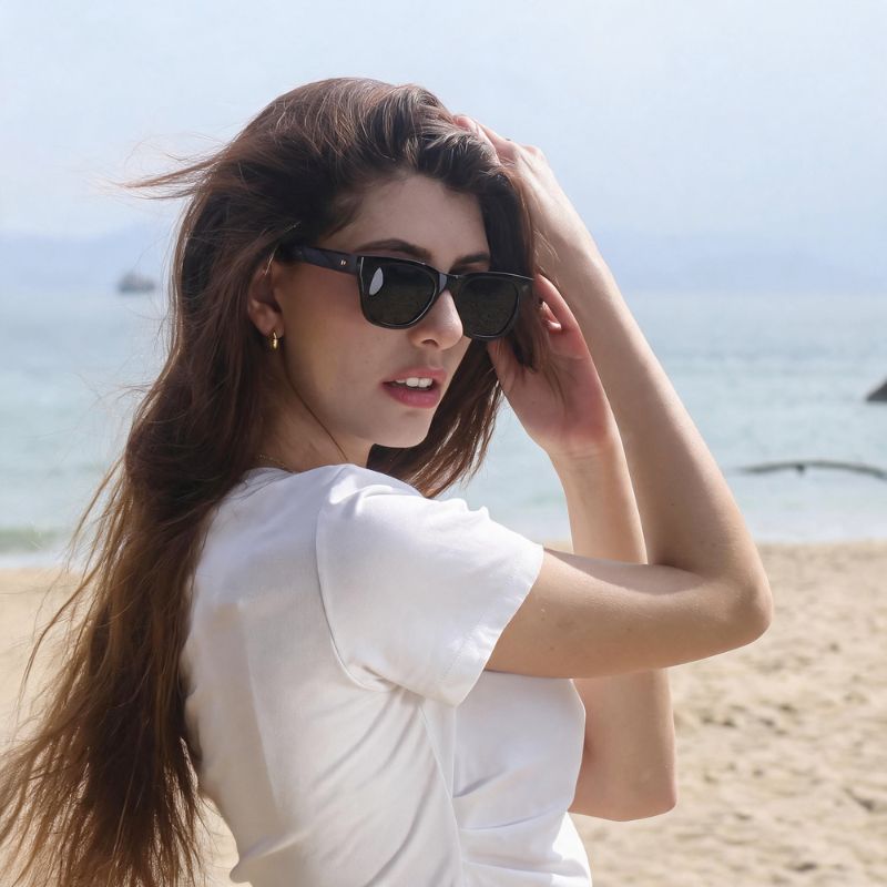 A woman with long brown hair stands on a sandy beach wearing BOBOTAILS matching dog harness leopard black cat-eye sunglasses and a white t-shirt, with the ocean and sky blurred in the background.