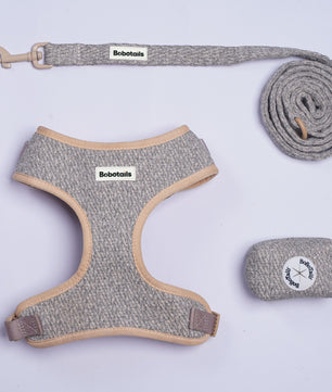 A chunky knit pet harness, leash, and storage pouch set by Bobotails, laid out on a light lavender background. The harness and leash are made of a heathered gray fabric with beige trim, and the leash has a beige metal clasp.