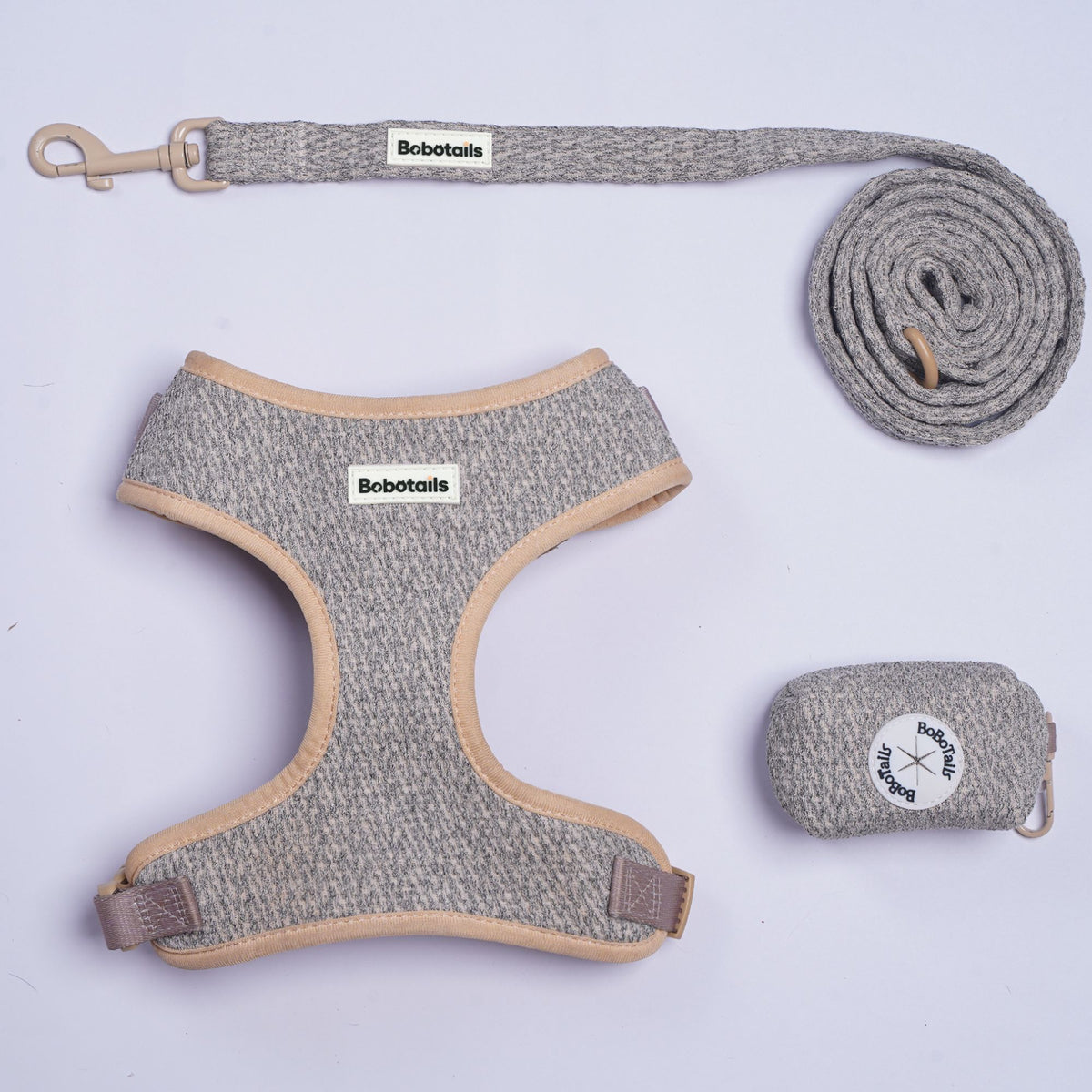 A chunky knit pet harness, leash, and storage pouch set by Bobotails, laid out on a light lavender background. The harness and leash are made of a heathered gray fabric with beige trim, and the leash has a beige metal clasp.