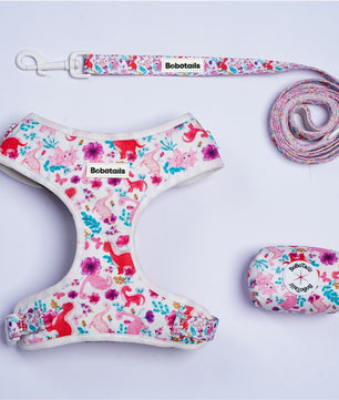 A pet accessory set laid out on a light background, including a floral-print harness, matching leash with clip, a coiled leash section, and a small zippered pouch. The fabric has a white base with pink, red, blue, and purple animal and floral print, and the brand name Bobotails is visible on the harness, leash, and pouch.