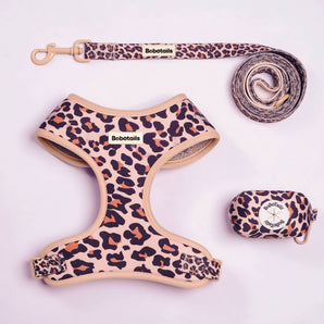 A pet gear set laid out on a light pink background, including a leopard-print harness, matching leash, and small matching pouch. The harness and leash have a beige base with black and brown leopard spots, and the brand name BoboTails is visible on the labels.