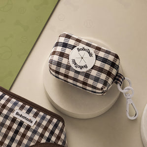 A small BoboTails zip pouch with a brown, beige, and black plaid pattern, featuring a white circular logo patch and a white carabiner clip, displayed on a beige surface.