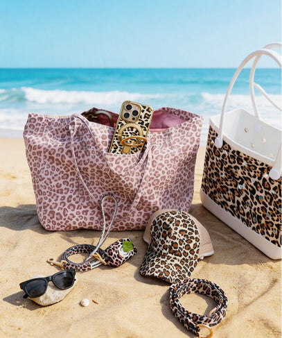 Leopard print accessories including a bag, hat, sunglasses, and bracelet on a sandy beach with ocean view.